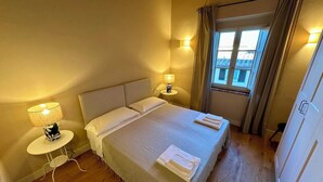 Una cama bien hecha con almohadas y una manta, una mesita de noche con lámpara, y una ventana con cortinas.