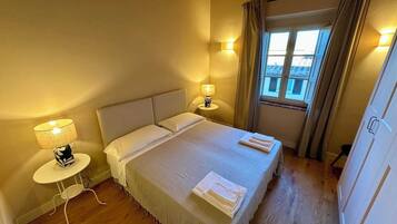 A neatly made bed with pillows and a blanket, a bedside table with a lamp, and a window with curtains.
