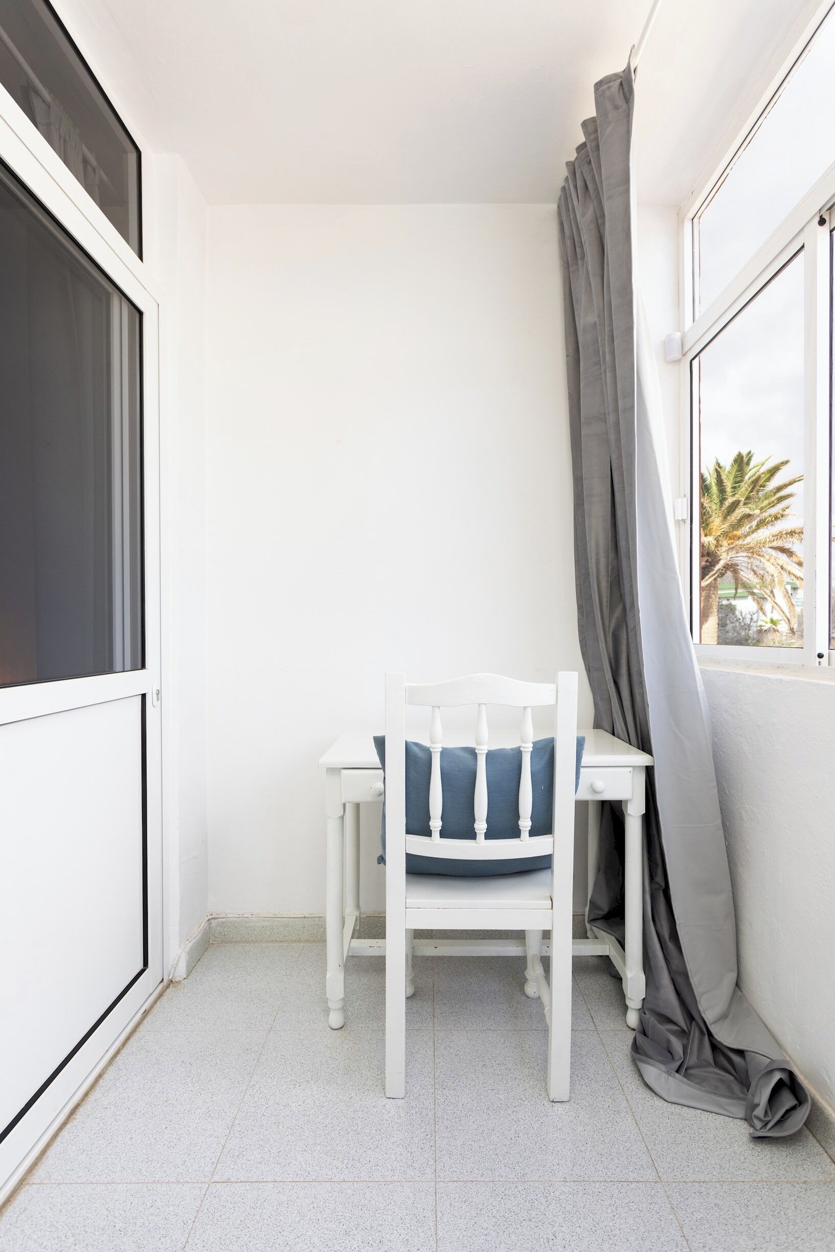 A small balcony with a white chair and table, a window with curtains, and a view of palm trees.