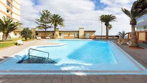 Una piscina con escalera metálica, rodeada de palmeras y sillas de descanso, con un edificio al fondo.
