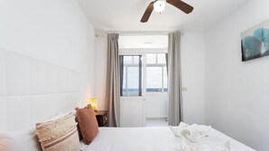 Habitación de hotel con cama, almohadas, mesita de noche con lámpara, ventilador de techo, ventana con cortinas y un cuadro en la pared.