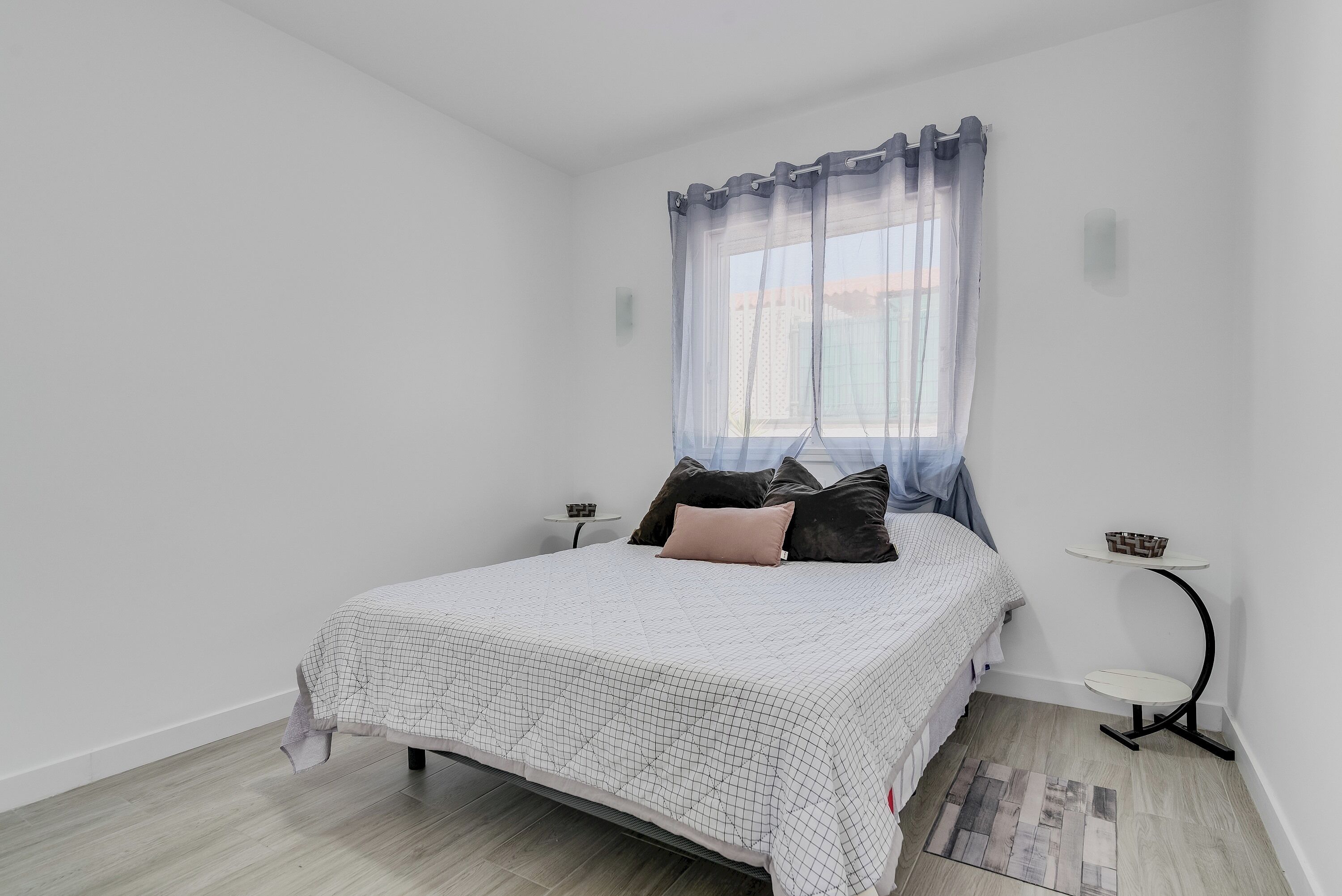 Un dormitorio con cama, mesita de noche, una pequeña mesa y una ventana con cortinas.