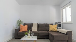 A modern living room with a sectional sofa, a small table with a bottle and glasses, a potted plant, and a window with an air conditioning unit. - Home2Book Vibrant Maspalomas Stay, Pool & Terrace (Maspalomas)