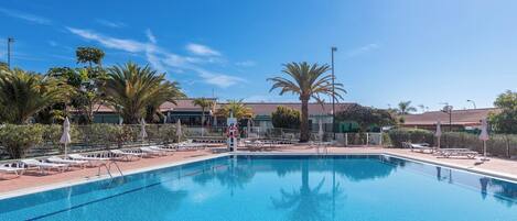 A clear blue pool with lounge chairs and umbrellas, surrounded by palm trees and a fence.