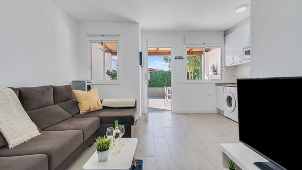 A modern living room with a sofa, coffee table, and TV. It leads to a kitchen with a washing machine and a dining area. - Home2Book Vibrant Maspalomas Stay, Pool & Terrace (Maspalomas)