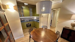 A dining area with a round wooden table and chairs, a hanging light fixture, and a view into a kitchen with green cabinets and a built-in oven. - Villa 16 (Bagno A Ripoli)