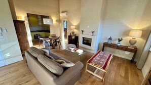 A spacious living area with a sofa, coffee table, and fireplace. A kitchen is visible in the background, and there is a dining table with chairs. - Villa 16 (Bagno A Ripoli)