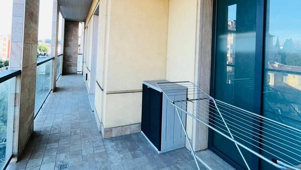 A corridor with a tiled floor, a metal railing, and a large window with a view of buildings.