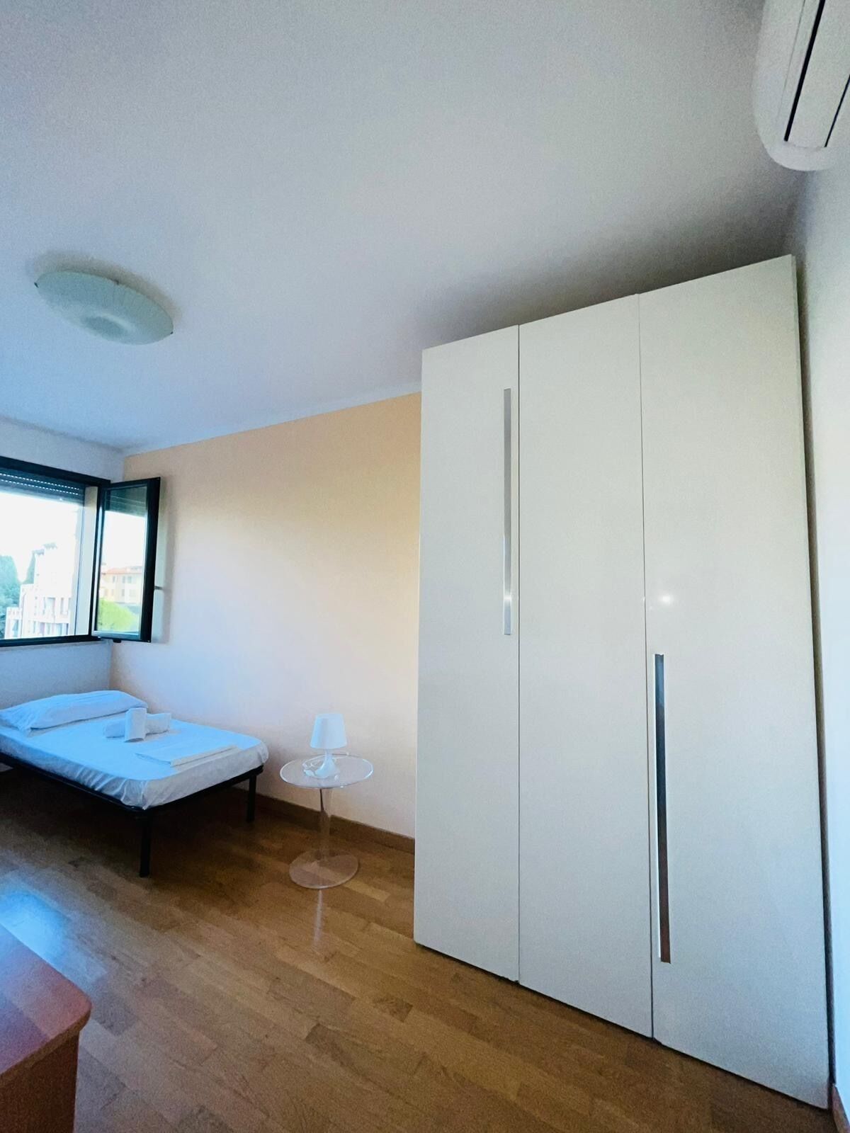 A bedroom with a bed, a small table, a wardrobe, and a window with a view of buildings.