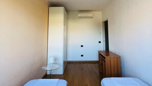 A hotel room with two single beds, a small table with a lamp, a wooden desk, and a wardrobe.