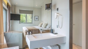 Une salle de bain moderne avec un grand lavabo blanc, un miroir, un sèche-cheveux et un téléviseur fixé au mur.