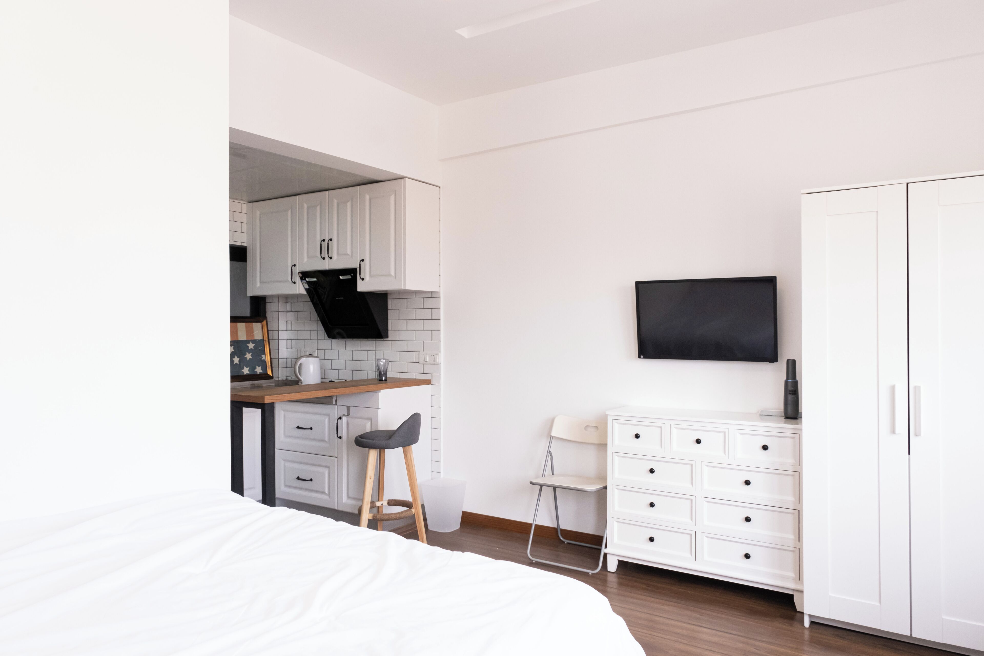 A modern bedroom with a large bed, a small kitchenette, and a TV mounted on the wall.