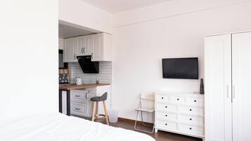 A modern bedroom with a large bed, a small kitchenette, and a TV mounted on the wall.