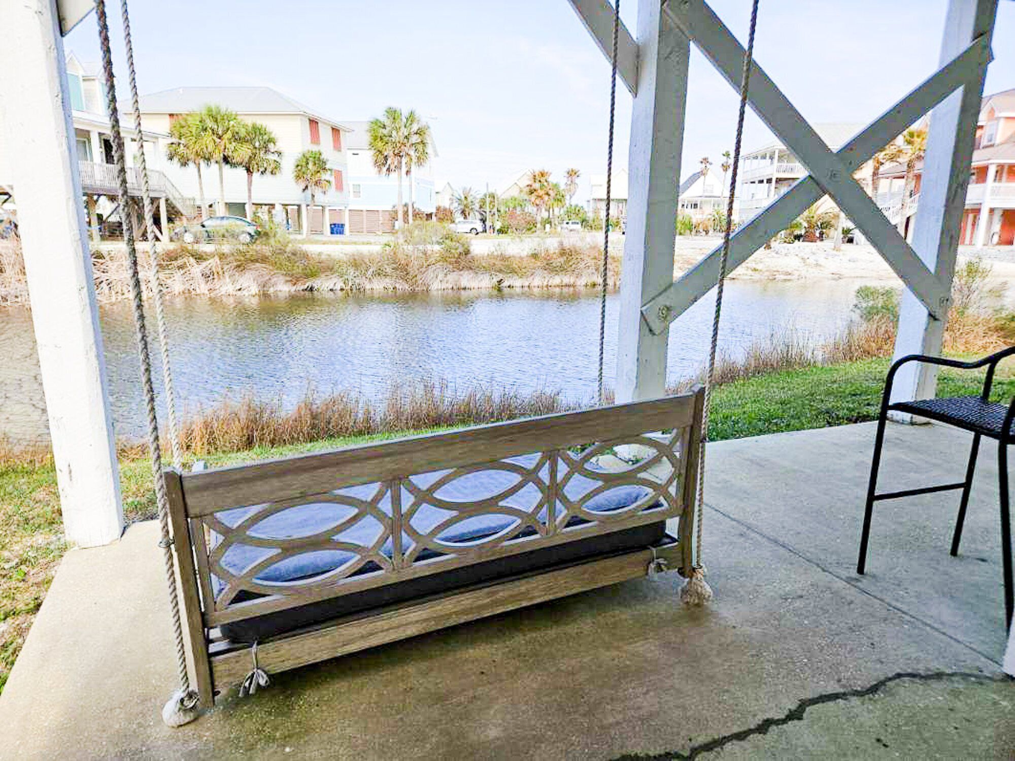 A wooden bench with a metal frame, positioned on a concrete surface near a water body, with a view of residential buildings in the background.