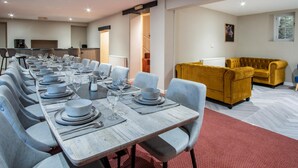 A dining area with a long table set for a meal, a yellow sofa, and a kitchen in the background. - The Lanterns - 9 Bedroom - Sleeps 17 - Saundersfoot Village Centre (Saundersfoot)