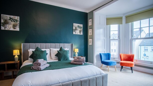 A bedroom with a large bed, two chairs, a window with a view, and artwork on the wall. - The Lanterns - 9 Bedroom - Sleeps 17 - Saundersfoot Village Centre (Saundersfoot)