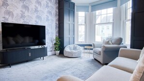 A modern living room with a large flat-screen TV, a dark blue accent wall, and a bay window with multiple windows. - The Lanterns - 9 Bedroom - Sleeps 17 - Saundersfoot Village Centre (Saundersfoot)
