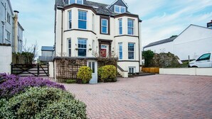A two-story house with a bay window, a front door, and a small garden with purple flowers. - The Lanterns - 9 Bedroom - Sleeps 17 - Saundersfoot Village Centre (Saundersfoot)