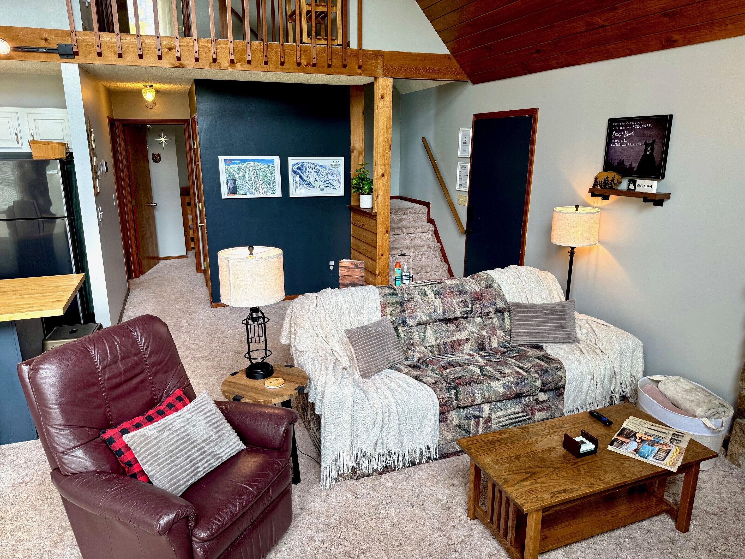 A cozy living room with a leather chair, patterned sofa, and a wooden coffee table. A staircase leads to an upper level, and a cityscape poster adorns the wall.