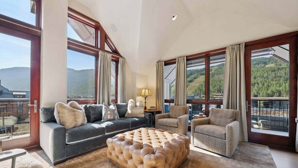 A spacious living room with a large tufted ottoman, a grey sofa with fur throw pillows, and a beige armchair. Large windows offer views of a mountainous landscape. - Solaris PHHW - 3 Bedroom (Vail)