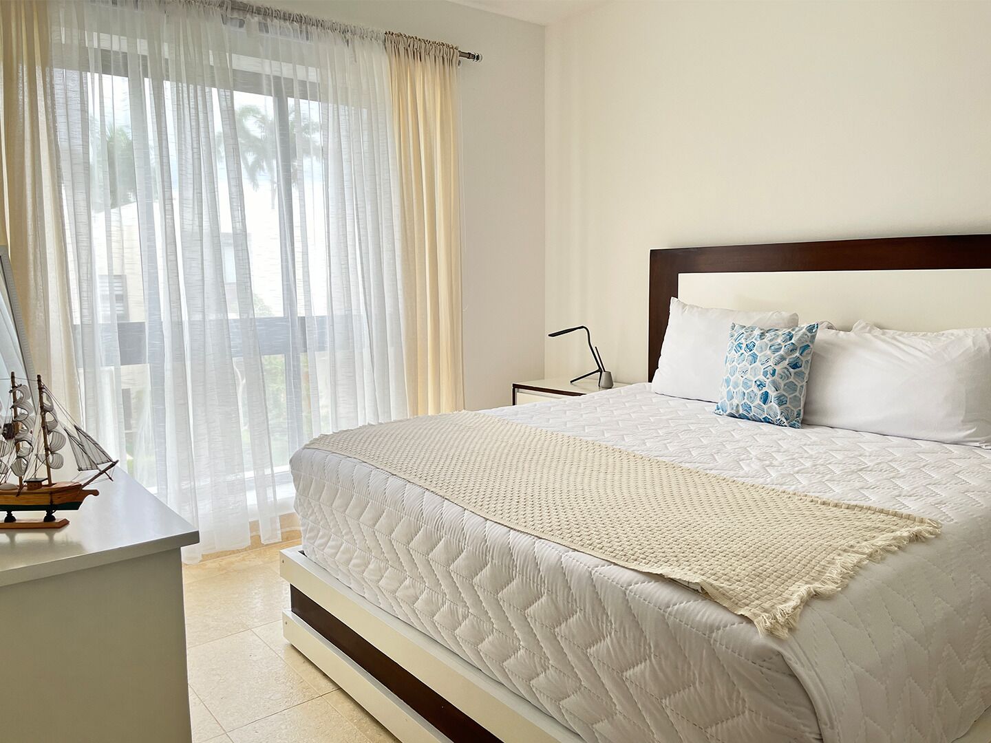 A bedroom with a large bed, white bedding, a wooden headboard, a nightstand, a lamp, and sheer curtains.