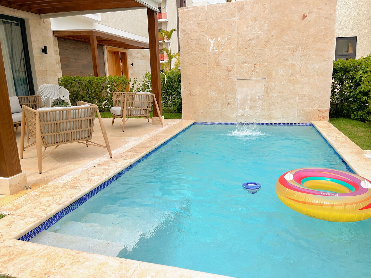 A rectangular swimming pool with a tiled edge, surrounded by a wooden pergola and wicker furniture.