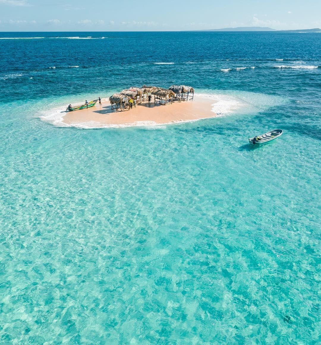 An aerial view of a small island with a thatched-roof structure surrounded by clear turquoise waters.