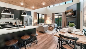 A modern kitchen with a central island, bar stools, and a dining area with a wooden table set for four. - Location! New Indoor POOL Cabin! Sunset Mnt View! Hot tub, Games, Bunk Room! (Sevierville)