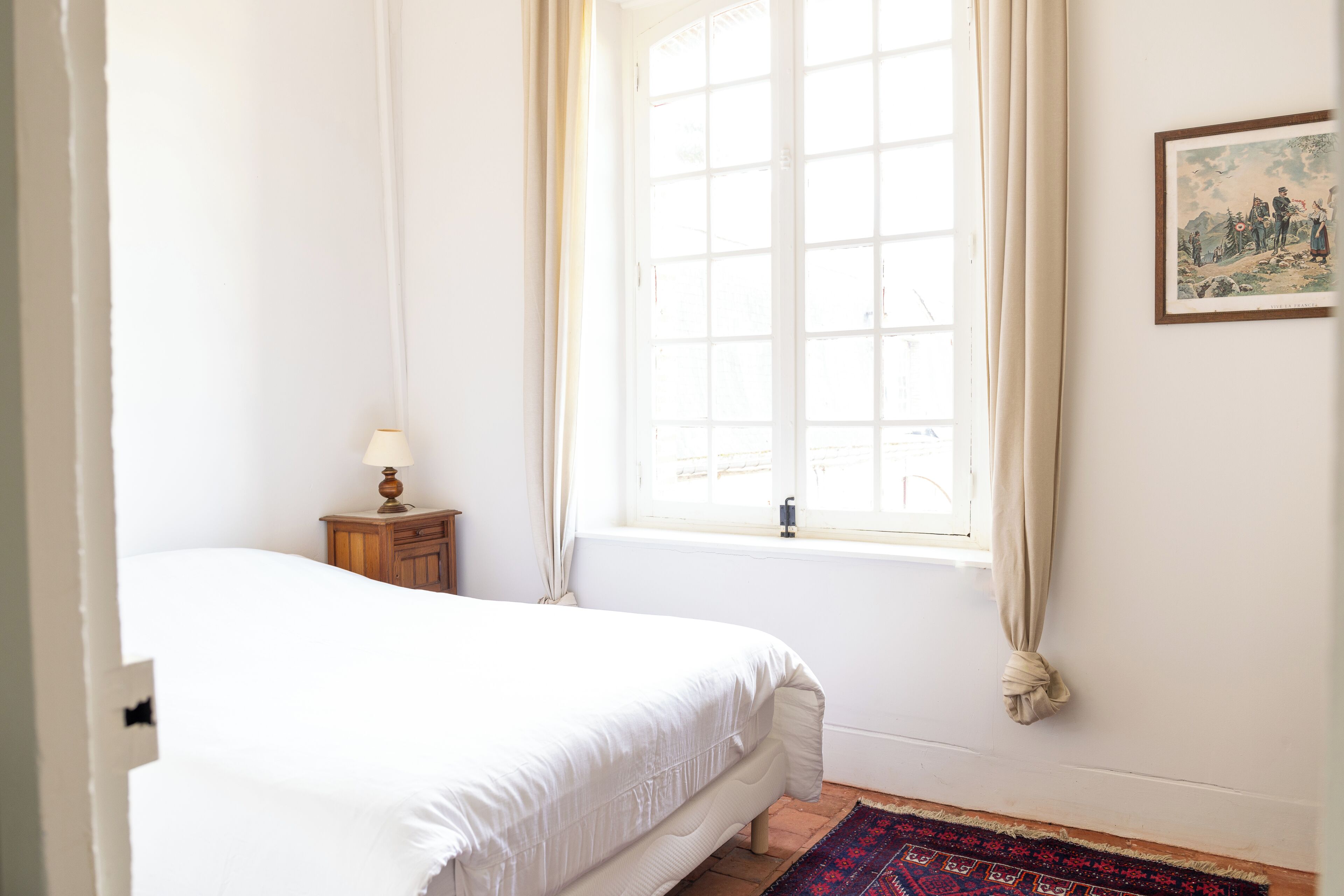 A bedroom with a large bed, a nightstand with a lamp, a window with curtains, and a framed picture on the wall.