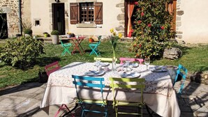 Un área de comedor al aire libre con dos mesas dispuestas para una comida, rodeada de muros de piedra y un edificio de piedra con contraventanas de madera.