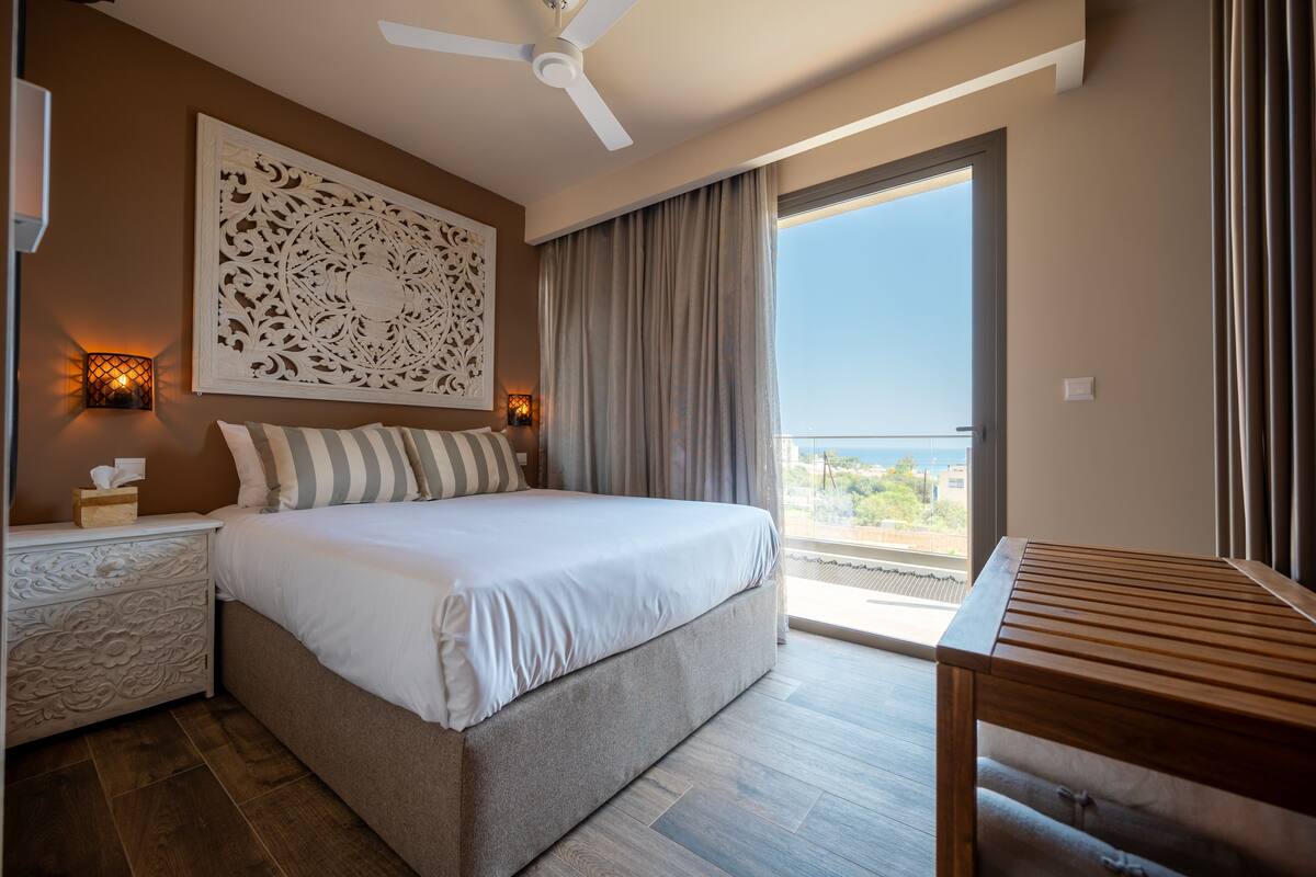 Ein Schlafzimmer mit Bett, Nachttischlampen, Deckenventilator, einem Holztisch und Blick ins Freie.