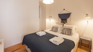 A neatly made bed with a dark blue bedspread, two folded white towels, and a hanging blue fabric on the wall. There are two wall-mounted lamps on either side of the bed.