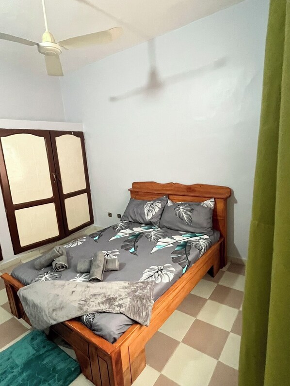 A single bed with a wooden frame, a grey floral bedspread, and a ceiling fan.