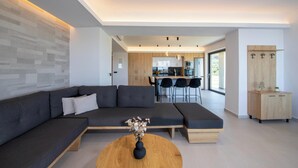 A modern living room with a sectional sofa, a round coffee table with a vase, and a kitchen in the background. - Villa Arithea (Chania)
