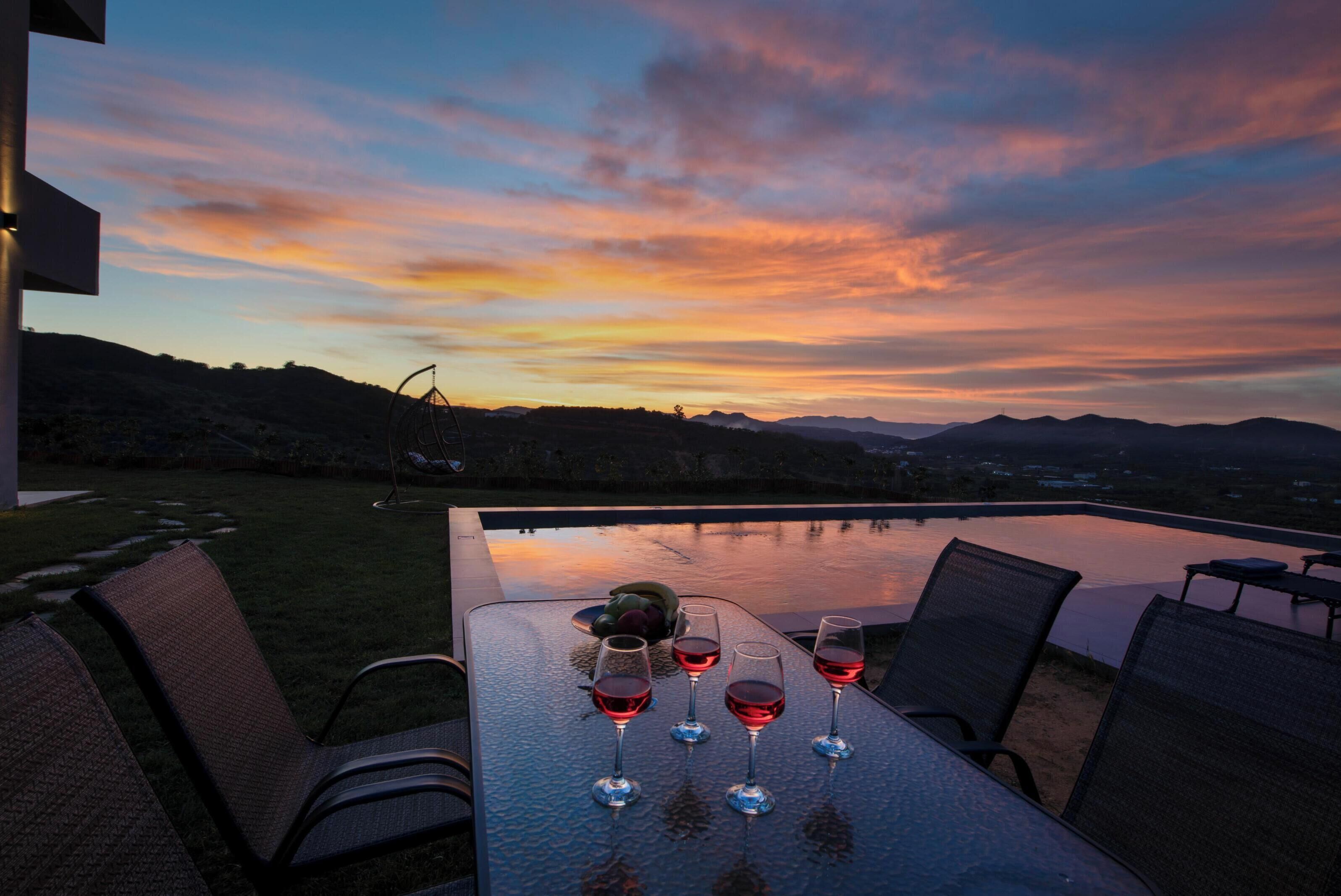 A sunset view from a patio table with four wine glasses and a bowl of fruit.