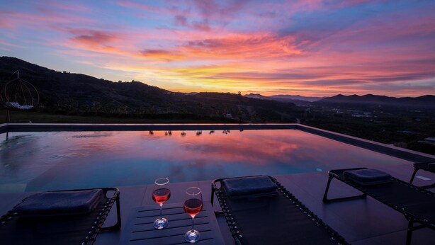 An infinity pool with lounge chairs and a glass of red wine, overlooking a mountainous landscape at sunset. - Villa Arithea (Chania)
