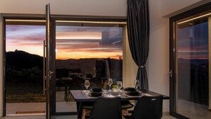 A dining area with a table set for two, a view of a sunset, and a sliding glass door leading to an outdoor area. - Villa Arithea (Chania)