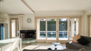 A modern living room with a sofa, a glass coffee table, a TV, and a view of a swimming pool through large sliding doors. - Villa Giuly (Sirmione)