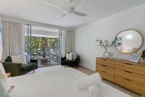 A hotel room with a bed, a wooden dresser, a round mirror, and a view of the outdoors.
