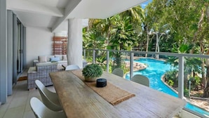 Uma varanda com mesa de madeira, mobiliário de vime e vista para uma piscina rodeada por vegetação tropical.