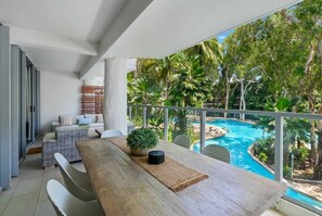 Un balcone con tavolo di legno, mobili in vimini e vista su una piscina circondata da vegetazione tropicale.