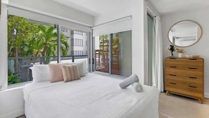 Quarto com uma cama grande, penteadeira de madeira, espelho redondo e porta de vidro deslizante para uma varanda com palmeiras.