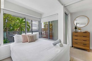 Una camera da letto con un grande letto, una cassettiera in legno, uno specchio rotondo e una porta scorrevole in vetro che dà su un balcone con palme.