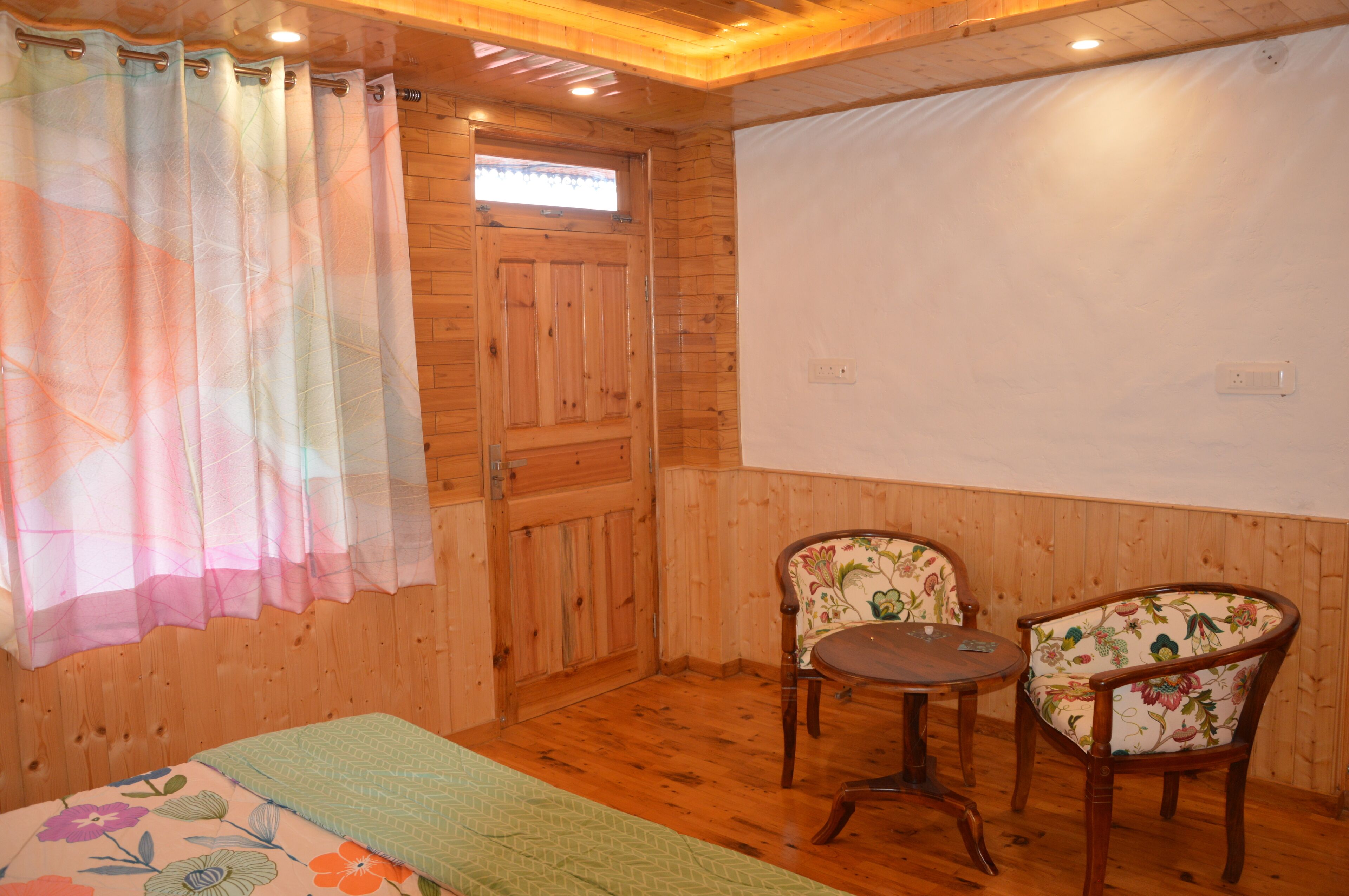 Una habitació amb terres de fusta, un llit, dues cadires, una taula petita i una finestra amb cortina.