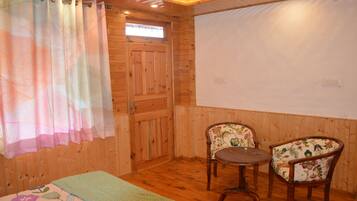 Una habitació amb terres de fusta, un llit, dues cadires, una taula petita i una finestra amb cortina.