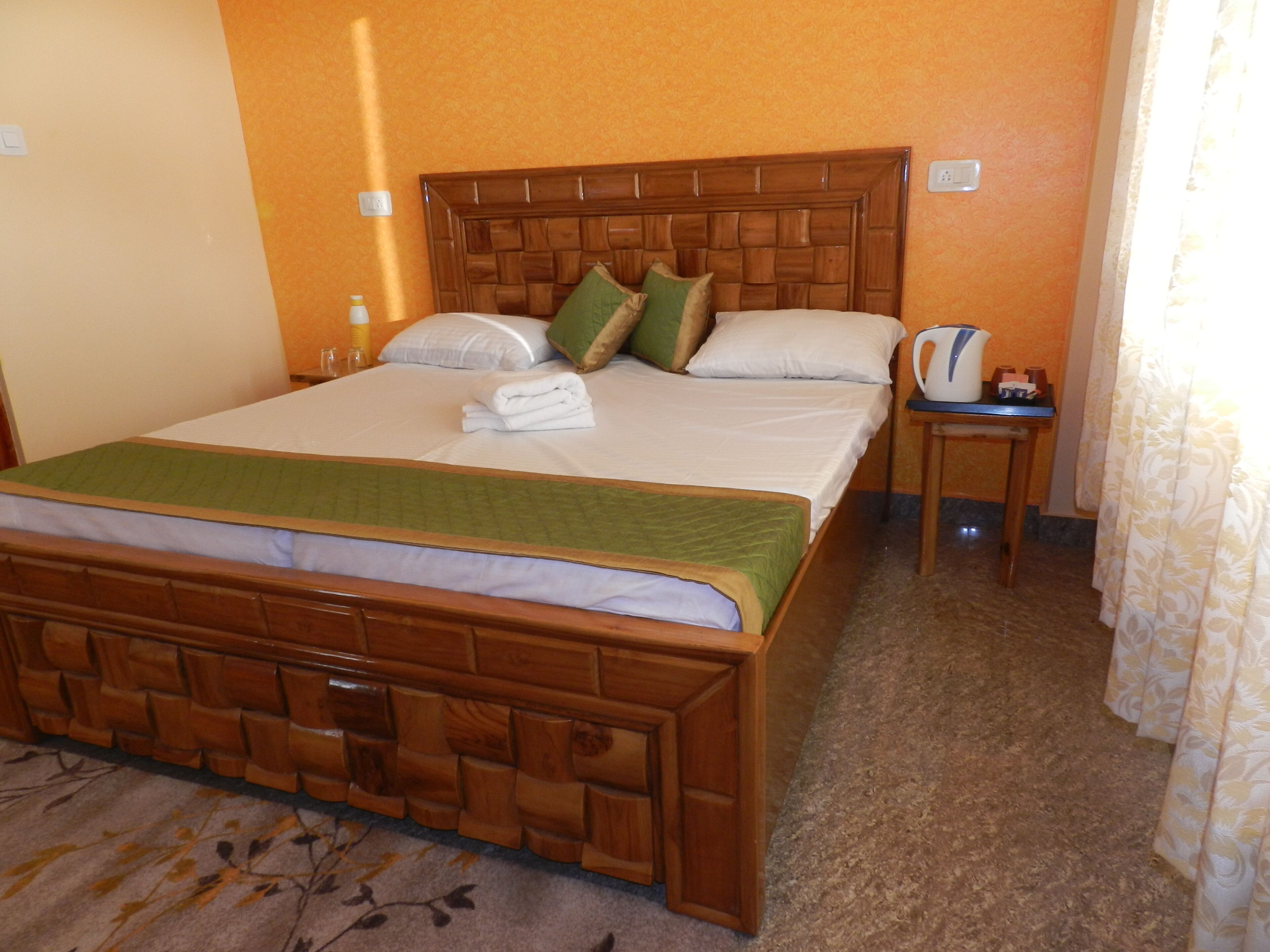 Un lit bien fait, avec une tête de lit en bois, une petite table de chevet sur laquelle se trouve une bouilloire, et un tapis à motifs floraux.