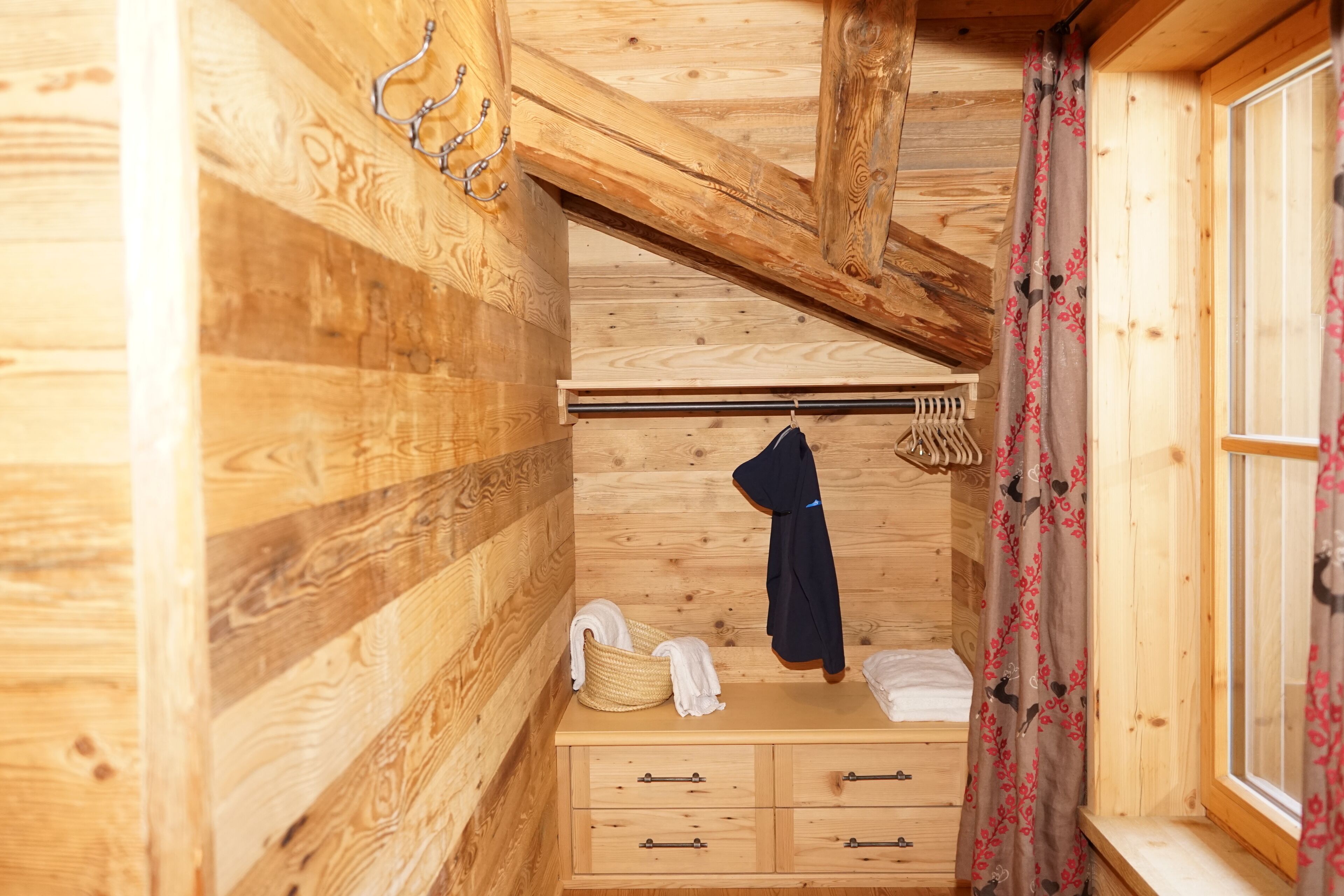 Interieur van een houten chalet met een houten dressoir, een kapstok en een raam met gordijnen.