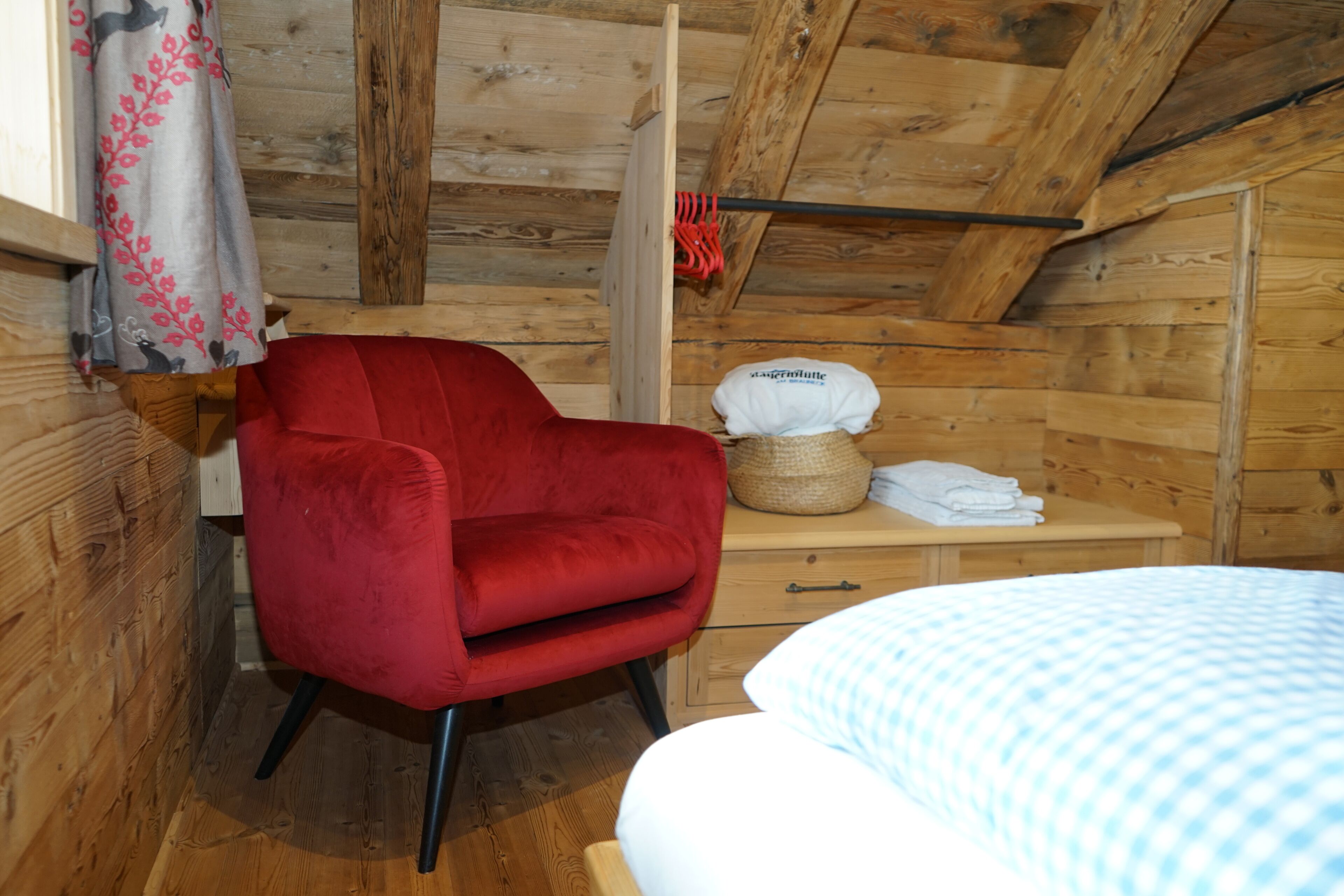 Ein Zimmer einer Holzhütte mit einem roten Sessel, einem Bett mit karierter Bettdecke und einer Holzkombination mit Korb und gefalteten Handtüchern.