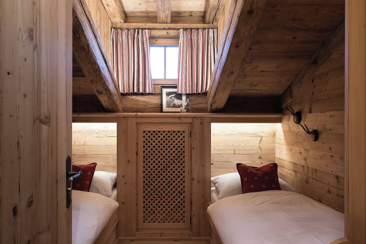 A wooden cabin room with two beds, a window with curtains, and a lattice partition.