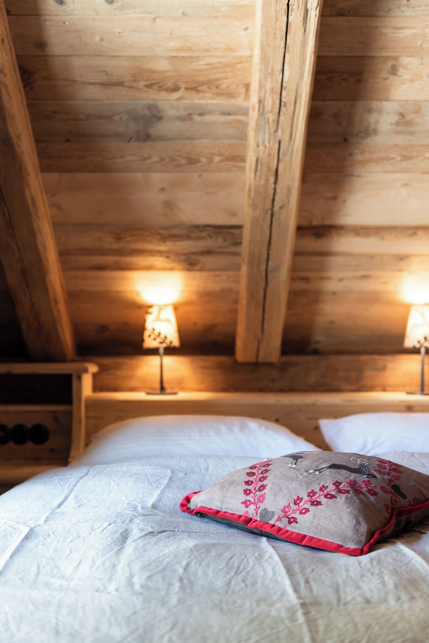 A wooden ceiling with exposed beams, a bed with white bedding and a decorative pillow, and two wall-mounted lamps.